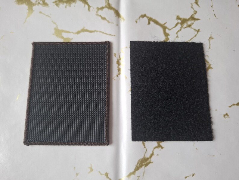 May include: Two square patches, one with a dark grey, textured surface and brown stitching, and the other with a black, fuzzy surface. The patches are displayed on a white background with gold accents.
