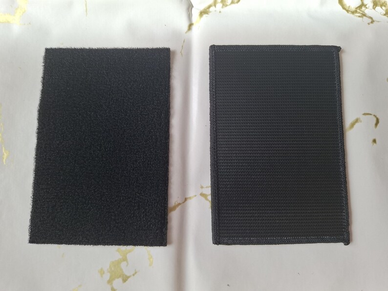 May include: Two black rectangular hook and loop fastener patches. One patch has a fuzzy surface, and the other has a woven texture with a stitched border. These are commonly used for attaching items.