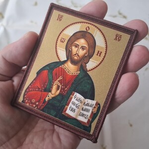 K&ouml;nnte beinhalten: Gestickter Aufn&auml;her mit einem religi&ouml;sen Bildnis von Jesus Christus. Das Bild zeigt Jesus mit Heiligenschein, der ein Buch mit Text h&auml;lt. Der Aufn&auml;her hat einen braunen Rand und einen goldenen Hintergrund. Die Ikone ist in Rot-, Gr&uuml;n- und Braunt&ouml;nen gehalten.