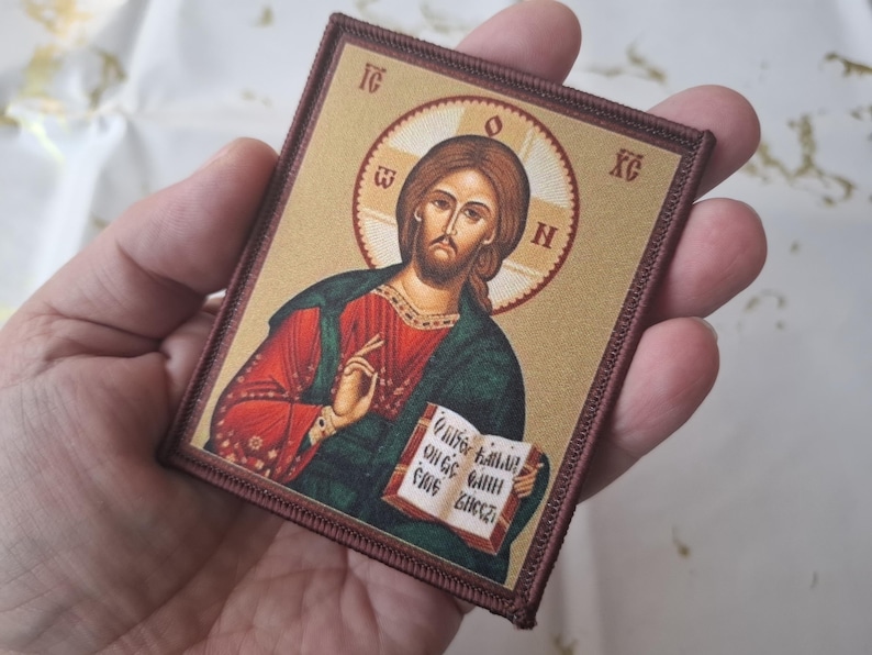 K&ouml;nnte beinhalten: Gestickter Aufn&auml;her mit einem religi&ouml;sen Bildnis von Jesus Christus. Das Bild zeigt Jesus mit Heiligenschein, der ein Buch mit Text h&auml;lt. Der Aufn&auml;her hat einen braunen Rand und einen goldenen Hintergrund. Die Ikone ist in Rot-, Gr&uuml;n- und Braunt&ouml;nen gehalten.