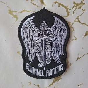 Könnte beinhalten: Schwarz-weißer, gestickter Aufnäher mit St. Michael, der ein Schwert hält und Flügel hat. Der Aufnäher hat den Text "ST. MICHAEL PROTECTUS" in weißer Schrift auf schwarzem Hintergrund. Oval geformt.