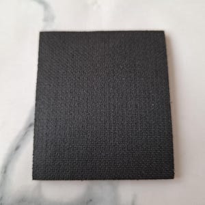May include: A square, black coaster with a textured surface. The coaster is made of a dark material and is set against a white background. The coaster is approximately 4 inches by 4 inches.