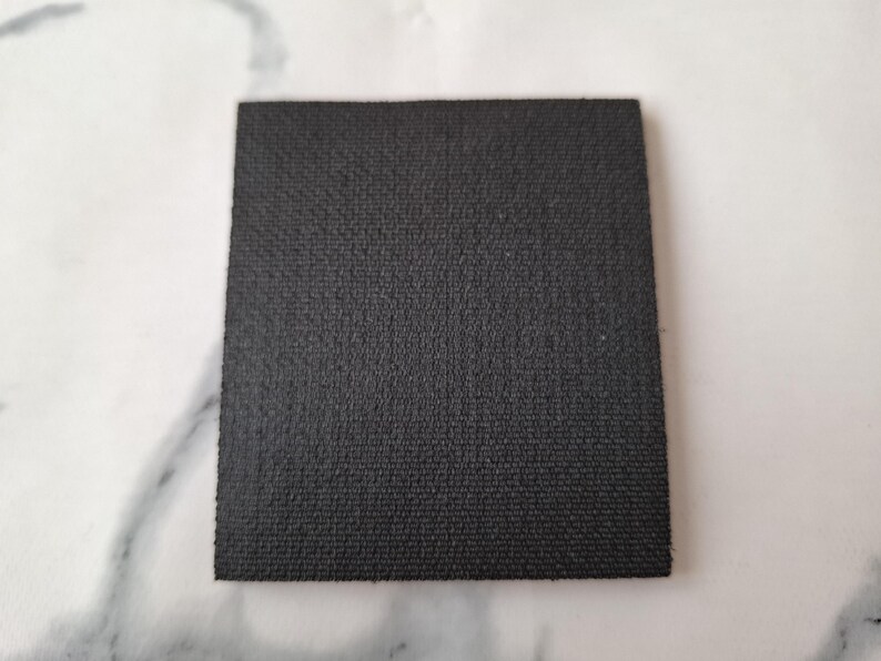 May include: A square, black coaster with a textured surface. The coaster is made of a dark material and is set against a white background. The coaster is approximately 4 inches by 4 inches.