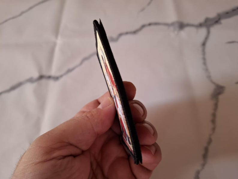 May include: A rectangular patch with a black border and a colourful design. The patch is held in a hand, showing its side profile. The design includes red, white, and other colours. The background is a white surface with grey veining.