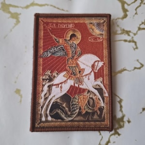 Może przedstawiać: Haftowana naszywka przedstawiająca ikonę religijną mężczyzny na białym koniu zabijającego węża. Naszywka ma czerwone tło ze złotymi akcentami i brązową obwódką. Tekst na obrazie jest w języku obcym.