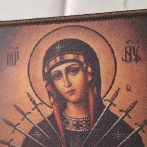 May include: Embroidered patch depicting a religious icon of a woman with a halo, wearing a crown and with several swords. The image has a vintage aesthetic with a brown border and detailed facial features.