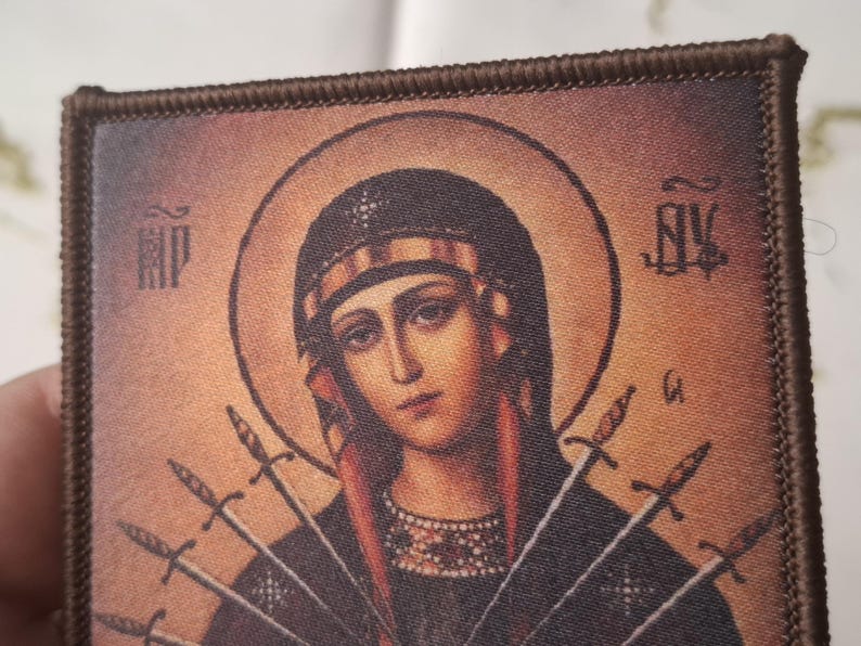 May include: Embroidered patch depicting a religious icon of a woman with a halo, wearing a crown and with several swords. The image has a vintage aesthetic with a brown border and detailed facial features.