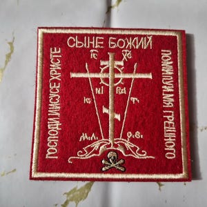Peut inclure: Écusson carré rouge avec texte brodé en or et un motif de croix. Le patch présente du texte cyrillique et une croix centrale avec des symboles supplémentaires. Un crâne et des os croisés sont en bas.