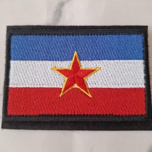 Könnte beinhalten: Gestickter Aufnäher mit der Flagge Jugoslawiens. Das Design zeigt horizontale Streifen in Blau, Weiß und Rot mit einem roten Stern in der Mitte. Der Aufnäher ist rechteckig mit einem schwarzen Rand.