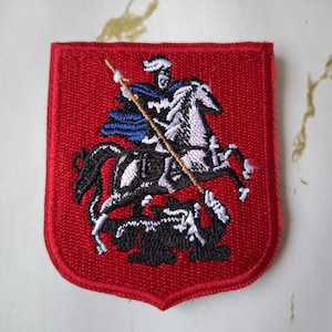 Könnte beinhalten: Gestickter Aufnäher mit einem Ritter auf einem Pferd, der einen Drachen ersticht. Das Design ist in Weiß, Schwarz und Blau auf einem roten, schildförmigen Hintergrund gehalten. Für Kleidung oder Accessoires.