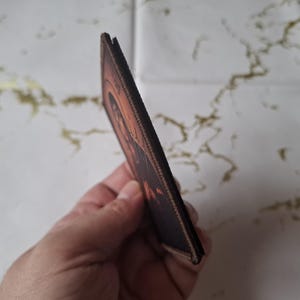 May include: A brown and orange rectangular patch featuring an image of a person. The patch has a stitched border and is held in a hand. The background is a white surface with gold markings.