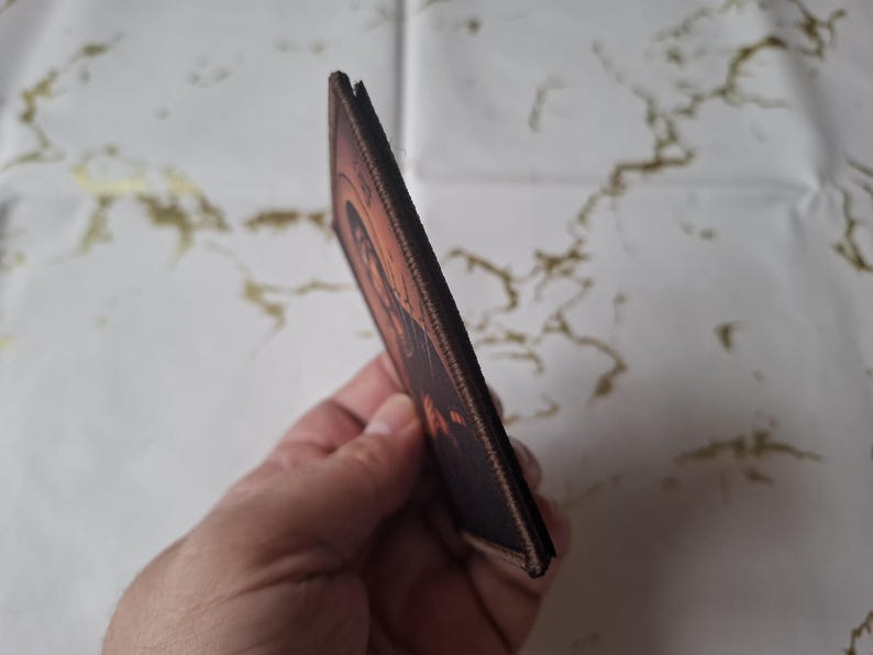 May include: A brown and orange rectangular patch featuring an image of a person. The patch has a stitched border and is held in a hand. The background is a white surface with gold markings.