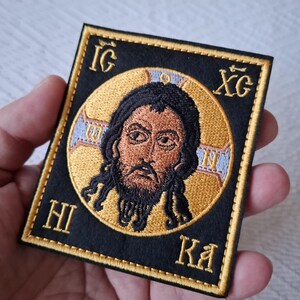 Op de afbeelding: Geborduurde patch met een religieus icoon van een man met een baard en lang haar, tegen een gouden halo. De afbeelding is omlijst door een zwarte rand met gouden Griekse letters. Een uniek religieus item.