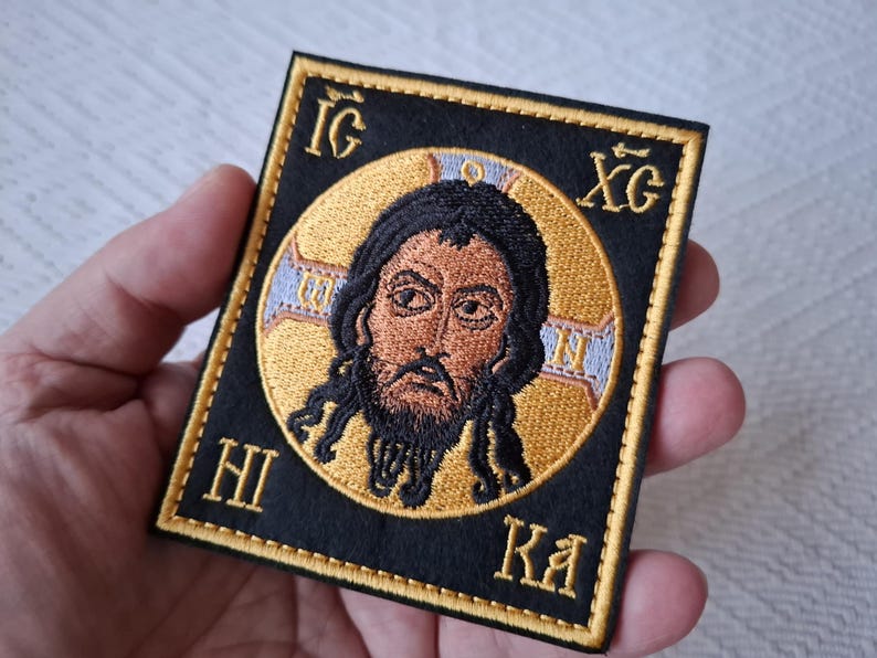 Op de afbeelding: Geborduurde patch met een religieus icoon van een man met een baard en lang haar, tegen een gouden halo. De afbeelding is omlijst door een zwarte rand met gouden Griekse letters. Een uniek religieus item.