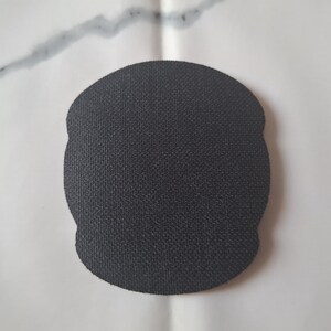 May include: A black, textured, oval-shaped coaster. The coaster has a slightly irregular edge and a woven appearance. The background is a white surface with a grey streak.