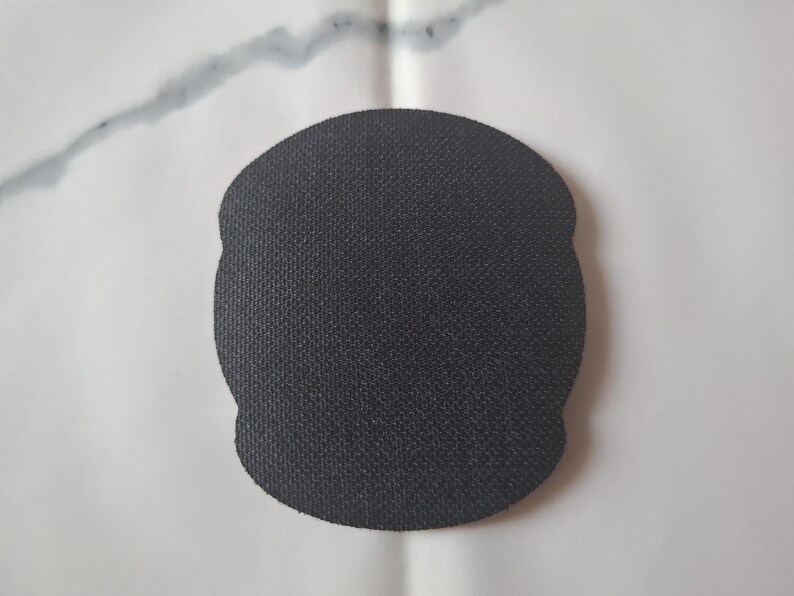 May include: A black, textured, oval-shaped coaster. The coaster has a slightly irregular edge and a woven appearance. The background is a white surface with a grey streak.