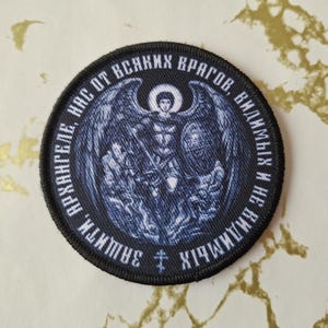 Peut inclure: Un écusson brodé noir et blanc représentant un ange avec des ailes déployées. Le texte autour de l'ange dit "АРХАНГЕЛЕ, ЗАЩИТИ НАС ОТ ВСЯКИХ ВРАГОВ, ВИДИМЫХ И НЕВИДИМЫХ".