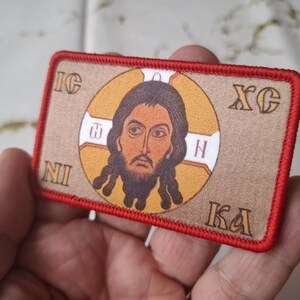Peut inclure: &Eacute;cusson rectangulaire avec une ic&ocirc;ne religieuse. L'image pr&eacute;sente un portrait central d'un homme barbu, entour&eacute; d'un cercle dor&eacute;. L'&eacute;cusson a une bordure rouge et du texte dans les coins.