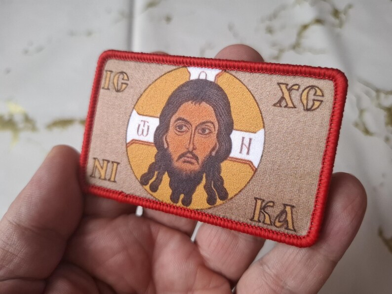 Peut inclure: &Eacute;cusson rectangulaire avec une ic&ocirc;ne religieuse. L'image pr&eacute;sente un portrait central d'un homme barbu, entour&eacute; d'un cercle dor&eacute;. L'&eacute;cusson a une bordure rouge et du texte dans les coins.
