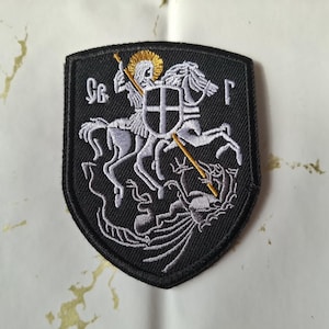 Puede incluir: Parche bordado negro que representa a un caballero a caballo matando a un dragón. El caballero, el caballo y el dragón están bordados en hilo blanco sobre un fondo negro en forma de escudo. Acentos dorados resaltan el halo y la lanza del caballero.