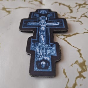 Pu&ograve; includere: Una croce ortodossa blu e nera con una dettagliata raffigurazione della crocifissione di Ges&ugrave; Cristo. La croce presenta disegni intricati e un teschio alla base, con testo nell'immagine.