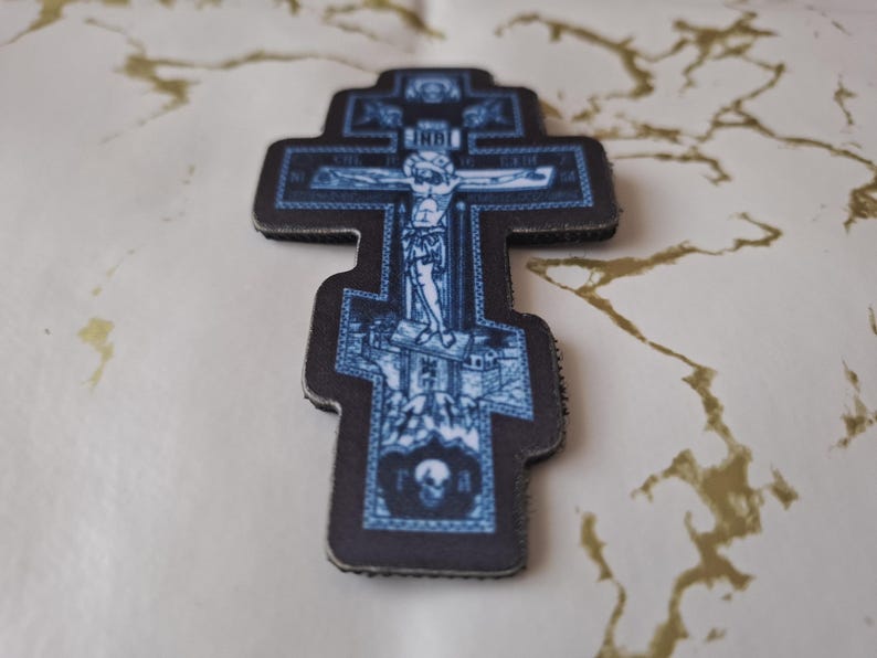 Pu&ograve; includere: Una croce ortodossa blu e nera con una dettagliata raffigurazione della crocifissione di Ges&ugrave; Cristo. La croce presenta disegni intricati e un teschio alla base, con testo nell'immagine.