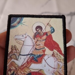 May include: Rectangular patch depicting a religious icon of a figure on horseback with a spear. The rider wears a red cloak and rides a white horse. The patch has a black border and a beige background.