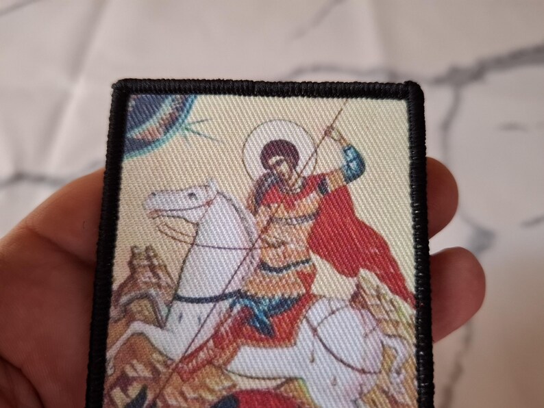 May include: Rectangular patch depicting a religious icon of a figure on horseback with a spear. The rider wears a red cloak and rides a white horse. The patch has a black border and a beige background.