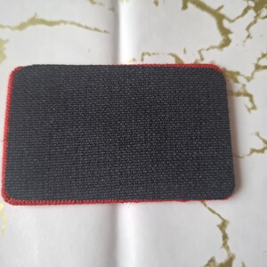 Peut inclure: Un patch rectangulaire noir avec une bordure rouge. Le patch a une surface textur&eacute;e et des coins arrondis. Le fond est blanc avec des marbrures dor&eacute;es.