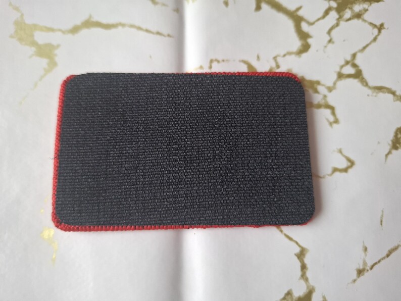 Peut inclure: Un patch rectangulaire noir avec une bordure rouge. Le patch a une surface textur&eacute;e et des coins arrondis. Le fond est blanc avec des marbrures dor&eacute;es.