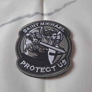 May include: Circular embroidered patch with a design of Saint Michael, holding a sword and shield. The patch has the words "Saint Michael" and "Protect Us" in white on a black and grey background. Suitable for application on clothing or accessories.