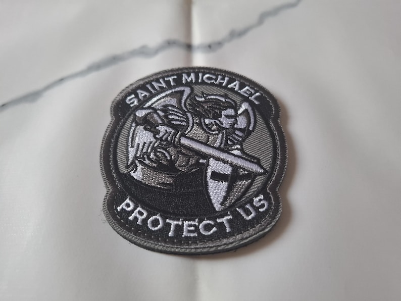 May include: Circular embroidered patch with a design of Saint Michael, holding a sword and shield. The patch has the words "Saint Michael" and "Protect Us" in white on a black and grey background. Suitable for application on clothing or accessories.