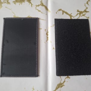 May include: Two rectangular black hook and loop fastener patches. One patch has a woven texture, and the other has a fuzzy texture. The patches are displayed on a white surface with gold accents.