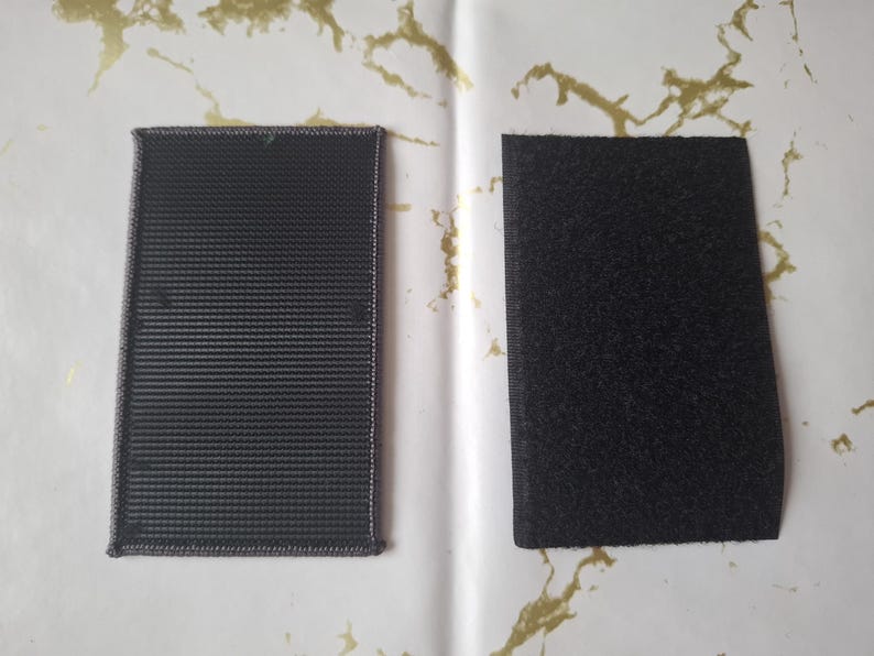 May include: Two rectangular black hook and loop fastener patches. One patch has a woven texture, and the other has a fuzzy texture. The patches are displayed on a white surface with gold accents.