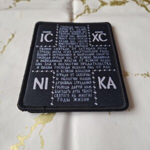 May include: Black rectangular patch with white text and symbols. The patch has a black border and is placed on a gold and white patterned surface. The text appears to be in a foreign language.