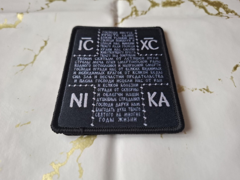 May include: Black rectangular patch with white text and symbols. The patch has a black border and is placed on a gold and white patterned surface. The text appears to be in a foreign language.