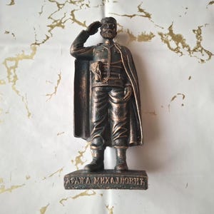 Puede incluir: Estatua de bronce de un hombre con uniforme militar, saludando. La figura lleva una capa y tiene bigote y barba. La estatua está montada sobre una base rectangular con texto cirílico. El fondo es blanco con vetas doradas.