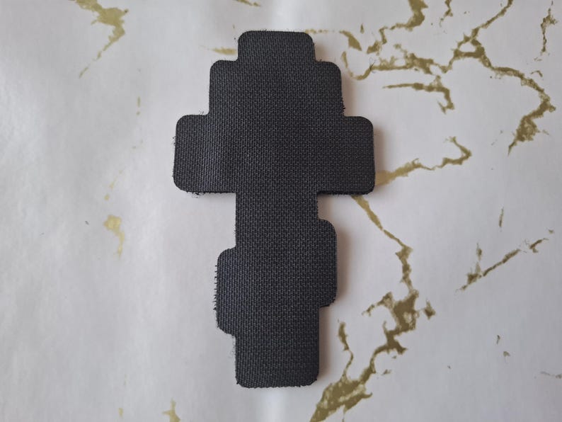 Pu&ograve; includere: Un oggetto nero, strutturato, a forma di croce. L'oggetto ha una serie di estensioni rettangolari. Lo sfondo &egrave; una superficie bianca con marmorizzazione dorata.