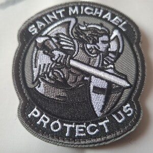 May include: Embroidered patch depicting Saint Michael, holding a sword and shield. The patch is predominantly grey and black, with white detailing. The text "SAINT MICHAEL PROTECT US" is embroidered around the image.