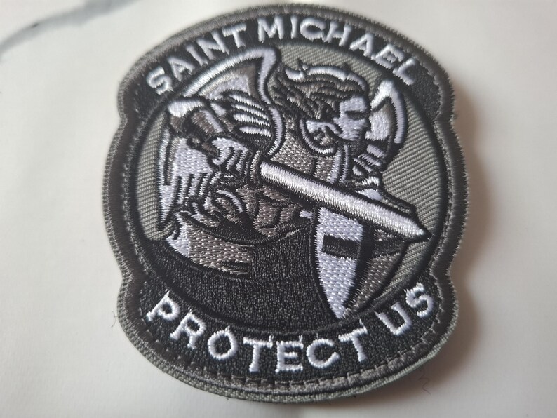 May include: Embroidered patch depicting Saint Michael, holding a sword and shield. The patch is predominantly grey and black, with white detailing. The text "SAINT MICHAEL PROTECT US" is embroidered around the image.