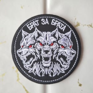 Könnte beinhalten: Ein runder, schwarzer, gestickter Aufnäher mit drei knurrenden Wolfsköpfen mit roten Augen und weißem Fell. Der Text "БРАТ ЗА БРАТА" ist in Weiß über den Wolfsköpfen angeordnet. Der Aufnäher hat einen gestickten Rand.