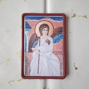 Könnte beinhalten: Rechteckiger Aufnäher mit einem religiösen Bildnis eines Engels mit braunem Haar und Flügeln. Der Engel trägt ein weißes Gewand und hält einen Stab. Das Bild hat einen roten Rand und ist vor einem Hintergrund aus Blau, Rosa und Braun gehalten.
