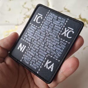 May include: Black rectangular patch with white text. The patch features religious text and symbols. The patch is held in a hand.