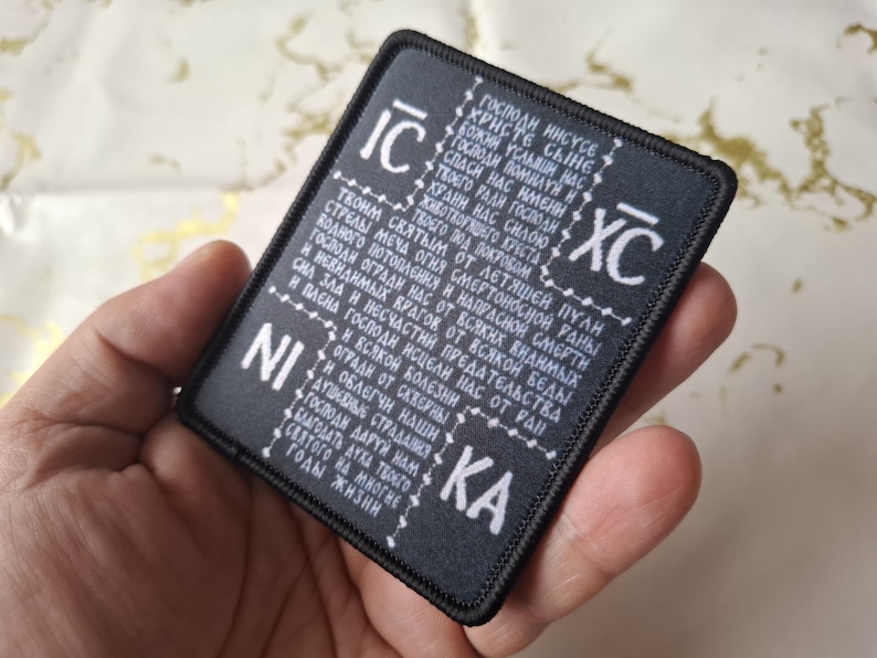 May include: Black rectangular patch with white text. The patch features religious text and symbols. The patch is held in a hand.