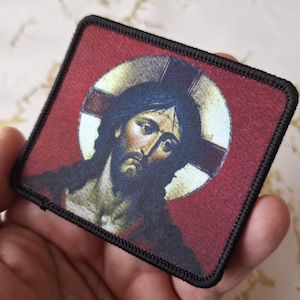 May include: A black and white embroidered patch with a red background featuring a depiction of Jesus Christ with a halo. The patch has a black border.