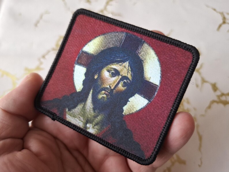 May include: A black and white embroidered patch with a red background featuring a depiction of Jesus Christ with a halo. The patch has a black border.