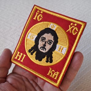 Op de afbeelding: Geborduurde patch met een religieus icoon. De patch heeft een rode achtergrond met gouden rand en letters. De centrale afbeelding is een afbeelding van een figuur met donker haar en een baard, geplaatst in een gouden cirkel.