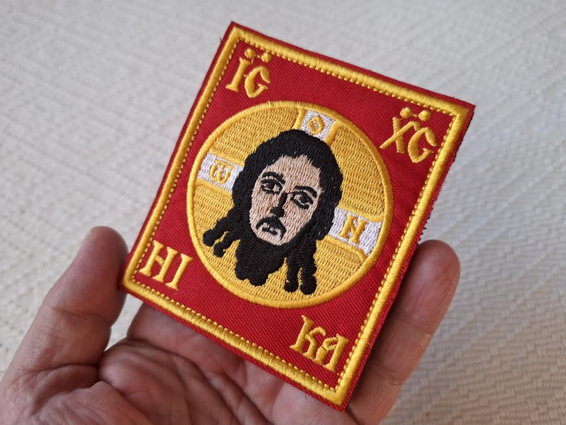 Op de afbeelding: Geborduurde patch met een religieus icoon. De patch heeft een rode achtergrond met gouden rand en letters. De centrale afbeelding is een afbeelding van een figuur met donker haar en een baard, geplaatst in een gouden cirkel.