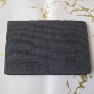 Peut inclure: Un &eacute;chantillon de tissu rectangulaire gris fonc&eacute; avec une surface textur&eacute;e. L'&eacute;chantillon est pr&eacute;sent&eacute; sur un fond blanc avec des marbrures dor&eacute;es. Le tissu semble &ecirc;tre tiss&eacute; serr&eacute;, sugg&eacute;rant une durabilit&eacute;.