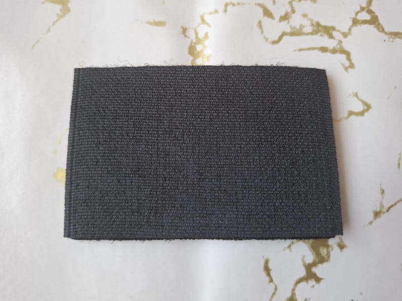 Peut inclure: Un &eacute;chantillon de tissu rectangulaire gris fonc&eacute; avec une surface textur&eacute;e. L'&eacute;chantillon est pr&eacute;sent&eacute; sur un fond blanc avec des marbrures dor&eacute;es. Le tissu semble &ecirc;tre tiss&eacute; serr&eacute;, sugg&eacute;rant une durabilit&eacute;.